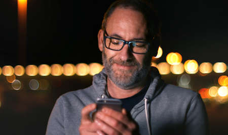 Handsome mature man speaking on smart phone at autumn sunset in city. Using smartphone for a phone call, smiling happy wearing urban hipster outfit outdoors at dawn.の写真素材