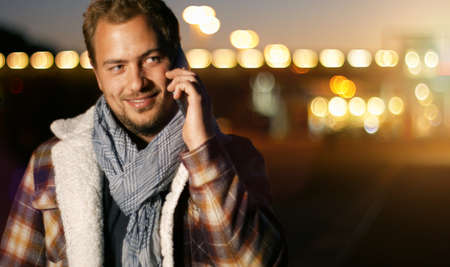 Handsome young man speaking on smart phone at autumn sunset in city. Using smartphone for a phone call, smiling happy wearing urban hipster outfit outdoors at dawn.の写真素材