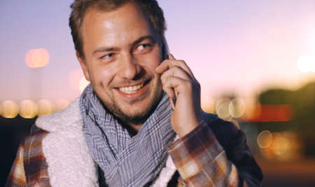 Handsome young man speaking on smart phone at autumn sunset in city. Using smartphone for a phone call, smiling happy wearing urban hipster outfit outdoors at dawn.の写真素材