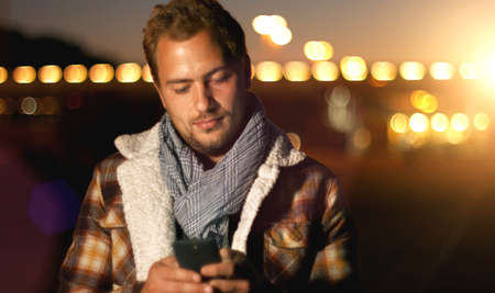Handsome young man sms texting using app on smart phone at autum Summer sunset in city. Using smartphone smiling happy wearing urban hipster outfit outdoors.の写真素材