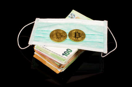 Bitcoin symbolic coin on the face mask and bunch of money banknotes isolated on the black background. Bitcoin over real money during corona virus financial crisis conceptの写真素材