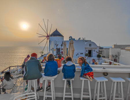 People sitting on chairs in restaurant in front of traditional Greek windmill in Oia village in Santorini, Greece in the late evening during a sunsetのeditorial素材