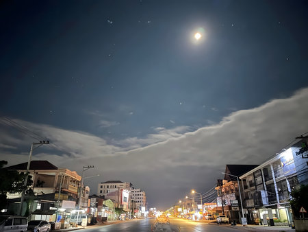 Quiet city street at night illuminated by street lights under a bright moon and cloudy sky, creating a calm and peaceful urban atmosphere.の写真素材