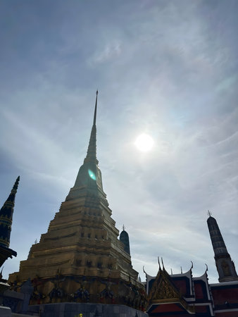 Low-angle view of an ancient golden pagoda at a traditional Buddhist temple, captured against a bright sky with sunlight.の写真素材