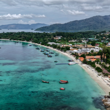 Aerial view of a tropical island coastline featuring turquoise sea, white sandy beach, coastal village, and traditional boats near the shore.の写真素材