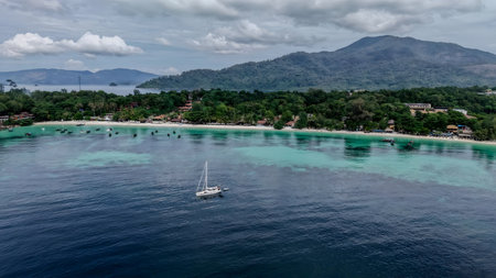 Aerial view of a tropical island coastline featuring a coastal village, white sandy beach, boats anchored along the shore, and clear turquoise sea.の写真素材