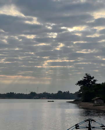 Soft sun rays breaking through dramatic clouds over a calm river, creating a peaceful and moody atmosphere. Natural landscape expressing hope, serenity, spirituality, and quiet reflection.の写真素材
