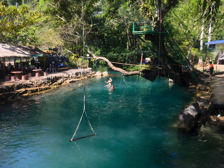 People enjoying a natural swimming pool with clear turquoise water and rope swing surrounded by lush tropical forest. Popular outdoor recreation and travel destination. Editorial use only.の写真素材