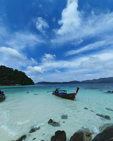 Tourists swimming and relaxing in shallow clear water near a traditional long tail boat at a tropical island beach. The scene shows real-life tourism activity with turquoise sea, rocky shoreline, island background, and dramatic blue sky during daytime.の写真素材