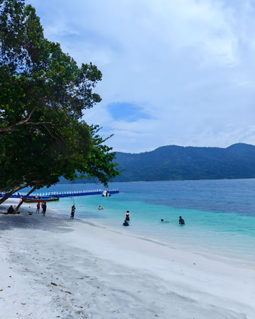 Tourists enjoying swimming and relaxing on a white sandy beach with clear turquoise sea and mountainous island background. The scene captures a peaceful tropical island atmosphere with people spending leisure time by the shoreline during daytime.の写真素材