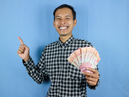 Excited expression handsome asian man hold paper money banknotes with pointing up gesture. Business...