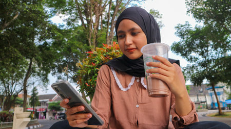 portrait of Muslim woman using mobile phone, reading or checking message at city park. Enjoying,...