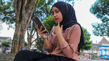 portrait of Muslim woman using mobile phone, reading or checking message at city park. Enjoying,...
