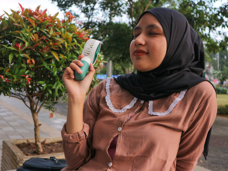 Portrait smiling of Asian muslim woman holding a portable electric fan near her face outdoors