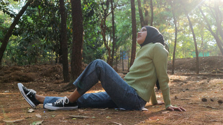 Young muslim woman resting carefree, relaxing on lawn on sunny day. Side view hipster woman with closed eyes enjoying calmness and freedom during coffee break while sitting on grass outdoors.の写真素材