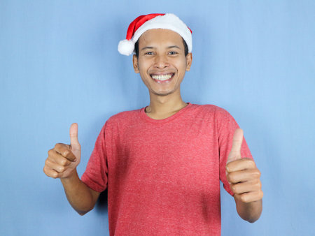 Handsome asian man in red christmas cap giving thumps up gesture isolated over blue background.の写真素材