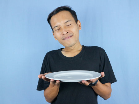 young Asian man in a black t-shirt stands and holds an empty white plate with the gesture of inhaling the dish.の写真素材