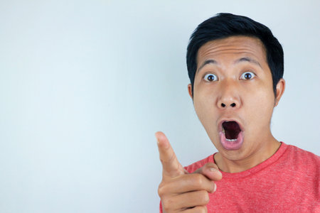 funny facial expression of asian man shocked and surprised while looking and pointing at the cameraの写真素材