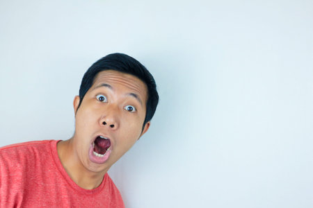 funny expression of shocked and surprised Asian man wearing a red t-shirt looking at the camera