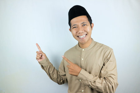 Smiling expression of muslim asian man, pointing finger an empty space. Advertising concept