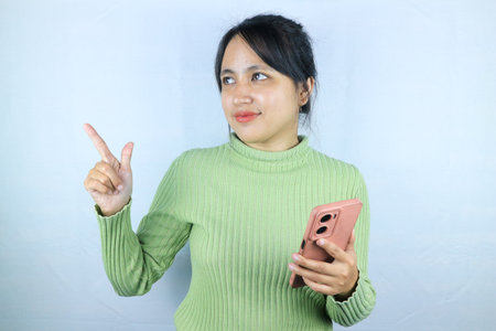 Smiling asian woman holding smart phone while pointing finger on white backgroundの写真素材
