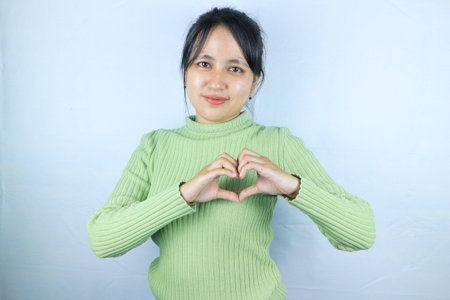 Smiling young asian woman making a heart gesture with her fingers in front of her chest showing her love and affection with a happy tenderの写真素材