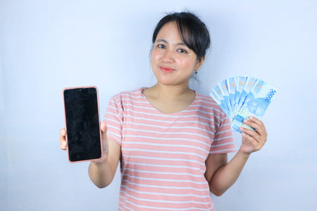 smiling expression of asian woman holding cash rupiah and showing empty mobile screen. Online...