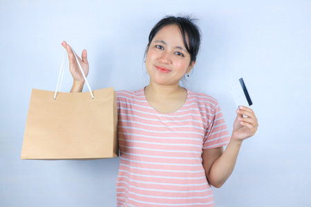 Young beautiful Asian woman holding paper shopping bag and showing credit card. Supermarket and...