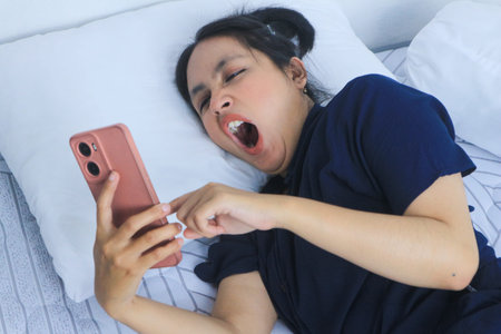 Tired beautiful Asian woman yawning in bed while holding phone. Bedtime concept