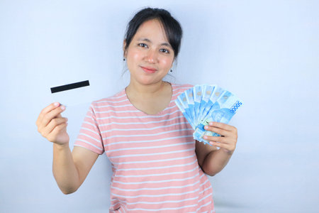 Portrait of a smiling asian young woman holding cash rupiah and credit card on white background
