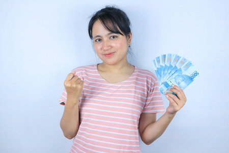 Portrait of a smiling asian young woman holding cash rupiah and celebrating on white background