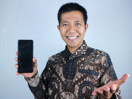 Smiling asian man wearing batik clothes showing smart phone with blank screen, mock up. white...