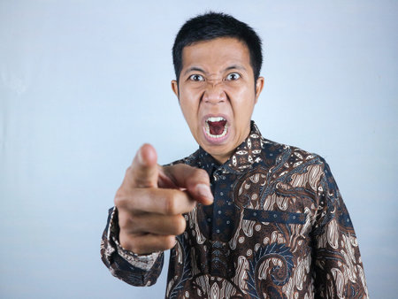 angry face expression of asian man wearing batik clothes while pointing at camera