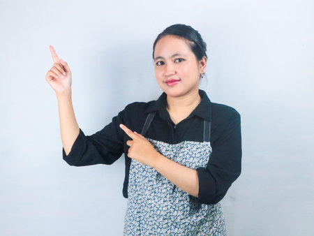 Smiling Asian woman in apron presenting or pointing finger with copy space, white backgroundの写真素材