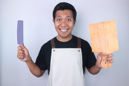 Young smiling asian men wearing apron hold knife and cutting board, preparing to cooking, look...