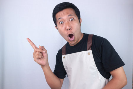 Funny shocked face expression young asian man wearing apron pointing right side, empty space. advertisement conceptの写真素材