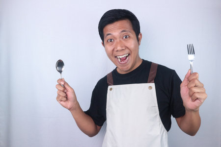 Hungry young Asian man wearing apron holding spoon and fork, feeling excited about eating delicious dish.の写真素材