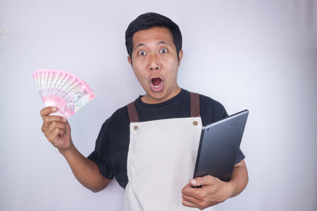 Funny surprise expression of asian man wear apron hold tablet and money. Small business concept