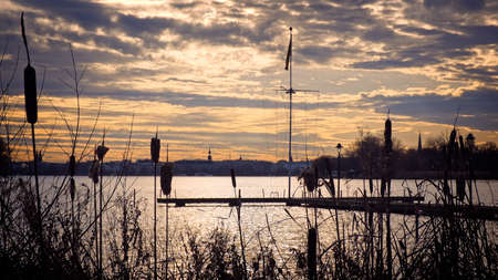 Bulrushes and wooden jetty silhouetted against a tranquil lake at sunset with colorful orange cloudy sky and reflection on the waterの写真素材