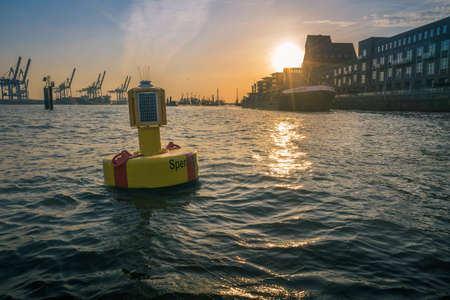 Floating buoy in a port with solar lantern to warn shipping of the channel boundaries with industrial cranes at a cargo depot silhouetted against a sunset and waterfront buildingsの写真素材