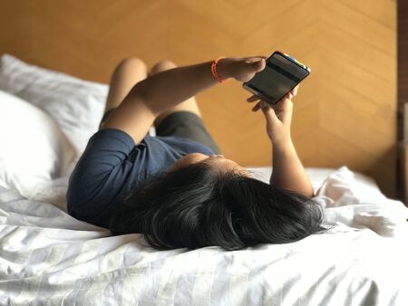 Asian woman wearing T shirt using smart phone on the white bed with her legs background in bedroomの写真素材
