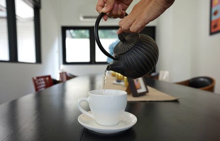 Human hands pouring the tea from black teapot into white porcelain ceramic tea cup in the cafeの写真素材