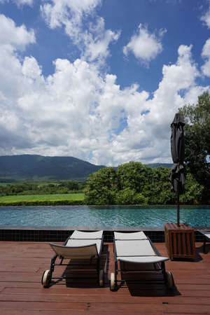 Recliner chairs on the deck next to swimming pool with green mountain and bright sky backgroundの写真素材