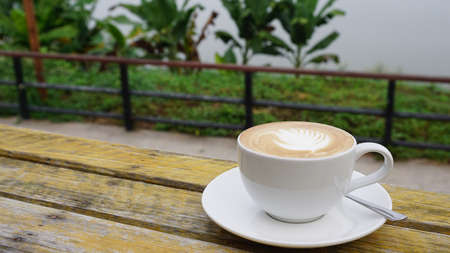 Hot latte coffee with leaf art in white cup on rustic timber table with nature backgroundの写真素材