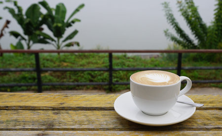 Hot latte coffee with leaf art in white cup on rustic timber table with nature background