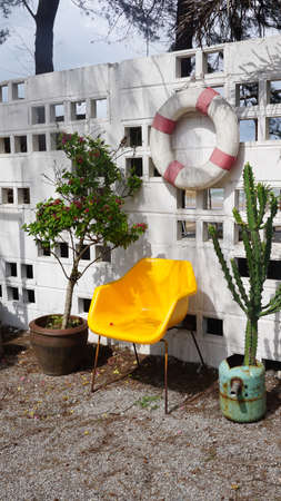 Yellow plastic chair with cactus and plants with lifebuoy hanging on white brick wall outdoorの写真素材