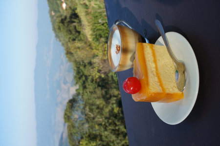 A cup of layer coffee in a glass cup with colorful orange layer cake and cherry on wooden table with...