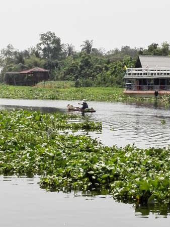 Landscape of river in Thailand with woman rowing and green trees and house on river bankの写真素材