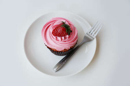 A photo of colorful cream strawberry cupcake decorated with strawberry topping and fork on white plate isolated on white background, close upの写真素材