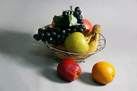 A photo of Artificial mixed fruits plastic, green and red apple, grape, orange, bananas on metal basket on gray background, close upの写真素材